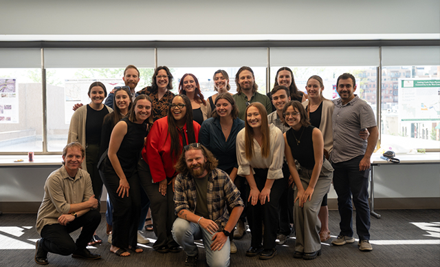 Students standing together and smiling at the Master of Urban and Regional Planning Capstone Celebration, while standing in front of windows taped with their posters from the semester. 