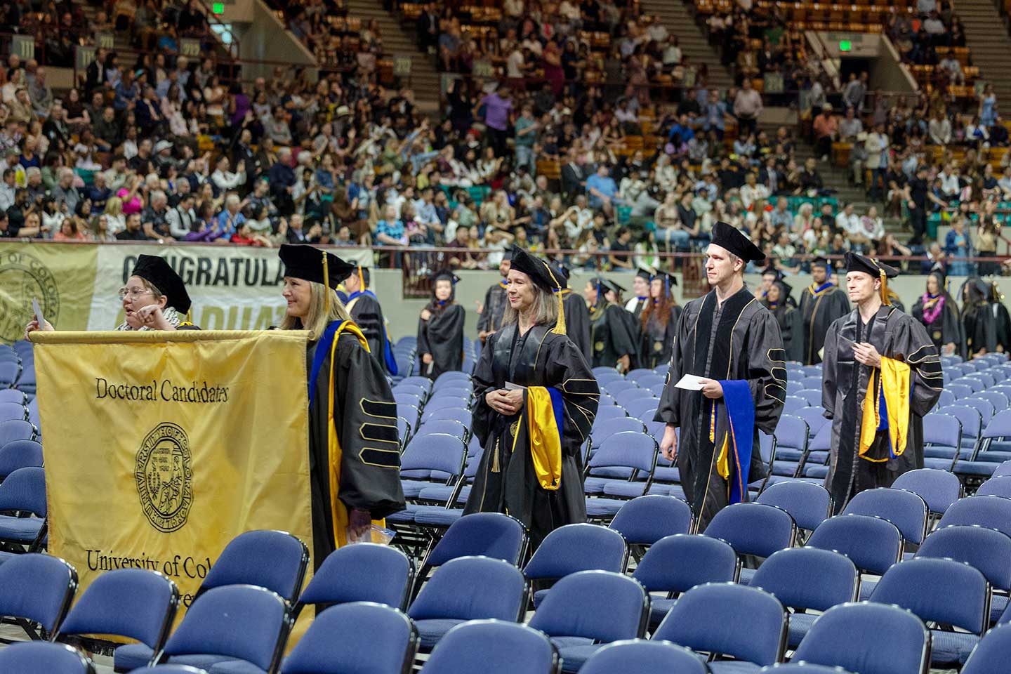 PhD students walk down an aisle at the Spring 2025 commencement ceremony carrying a Doctoral Candidate banner.