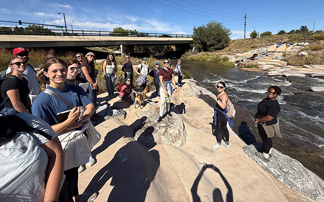 A group of students standing along a river near an overpass.