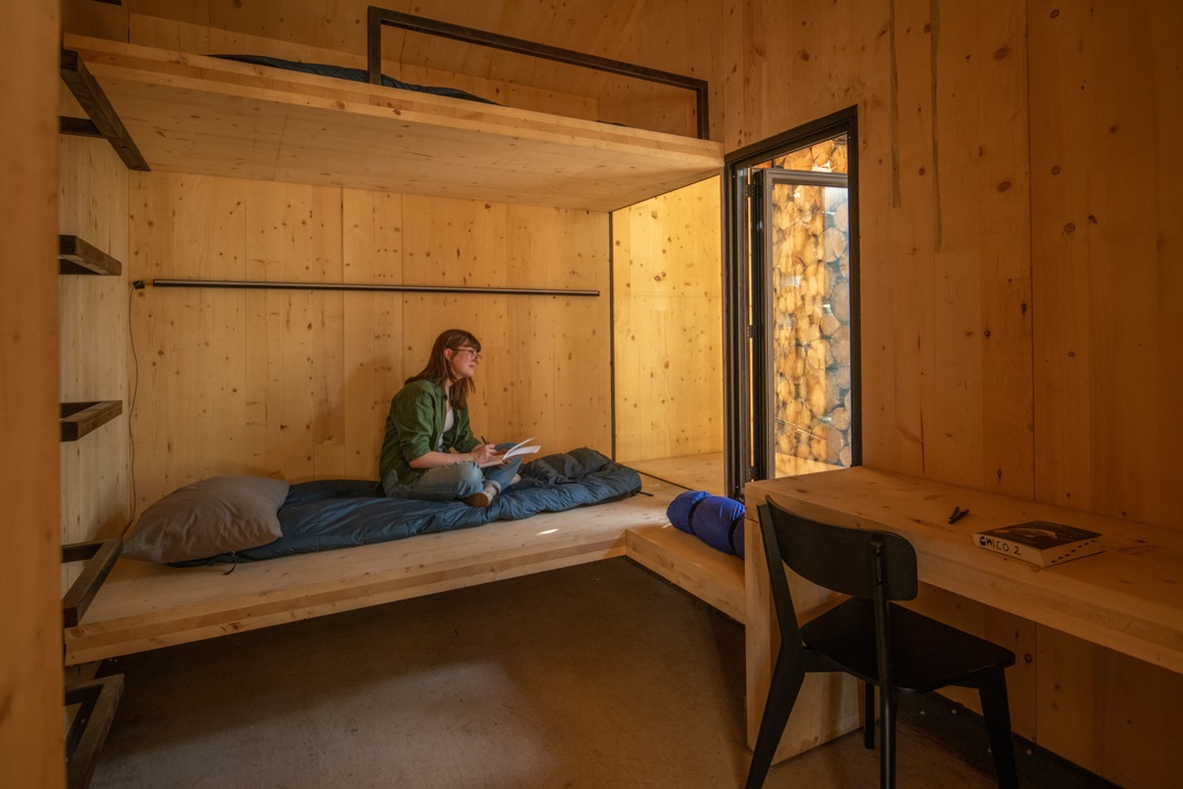 A student sitting on a sleeping bag while looking out the window and sketching in the cabin's dorm